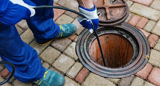A worker uses a cable to clean a sewer drain, surrounded by paving stones.