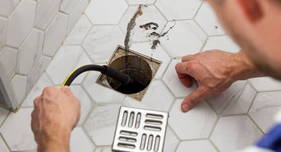 A person using a plumbing snake to unclog a floor drain on hexagonal tiled flooring.