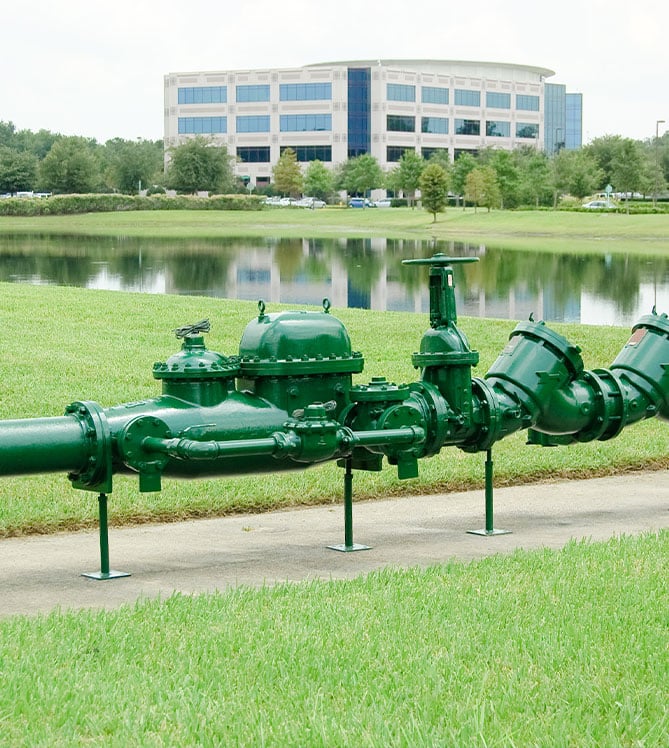 Green backflow preventer device near pond and office building in the background.