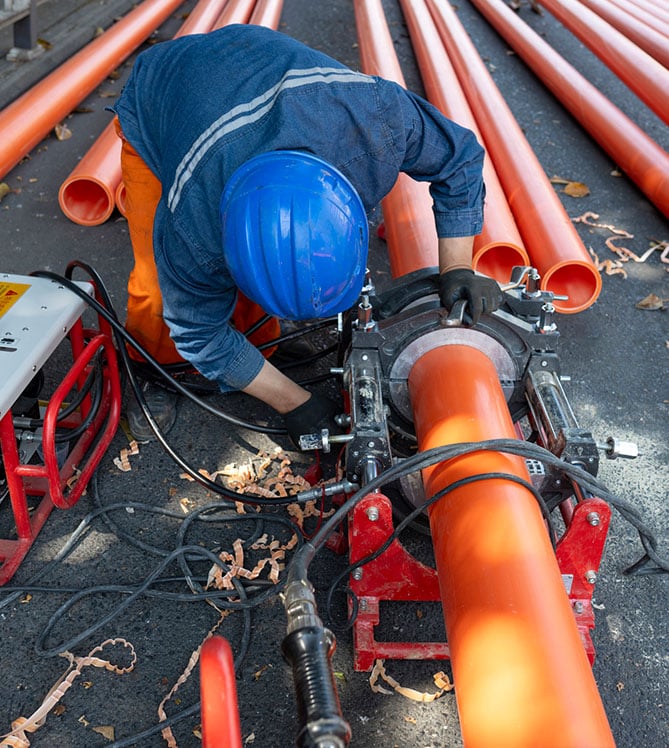 A worker wearing a blue helmet welding large orange plastic pipes outdoors.