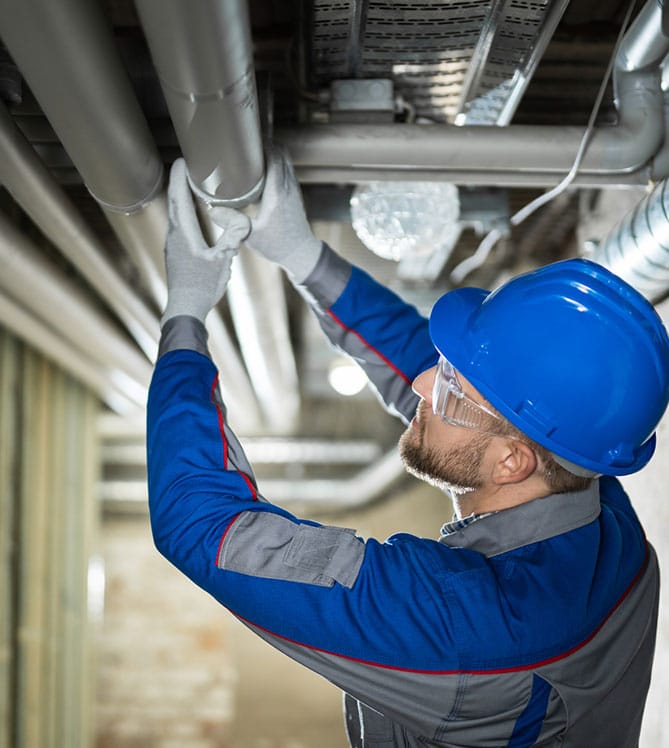 Worker in blue uniform and hard hat adjusting ceiling pipes.