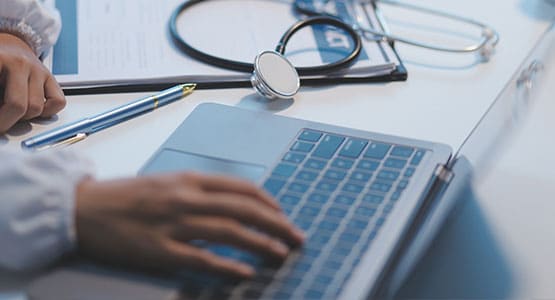 A person typing on a laptop with a stethoscope and clipboard on the desk.