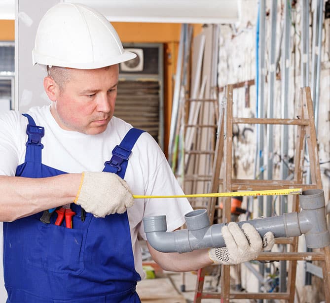 Worker in blue overalls and hard hat measuring a pipe with a tape measure on a construction site.
