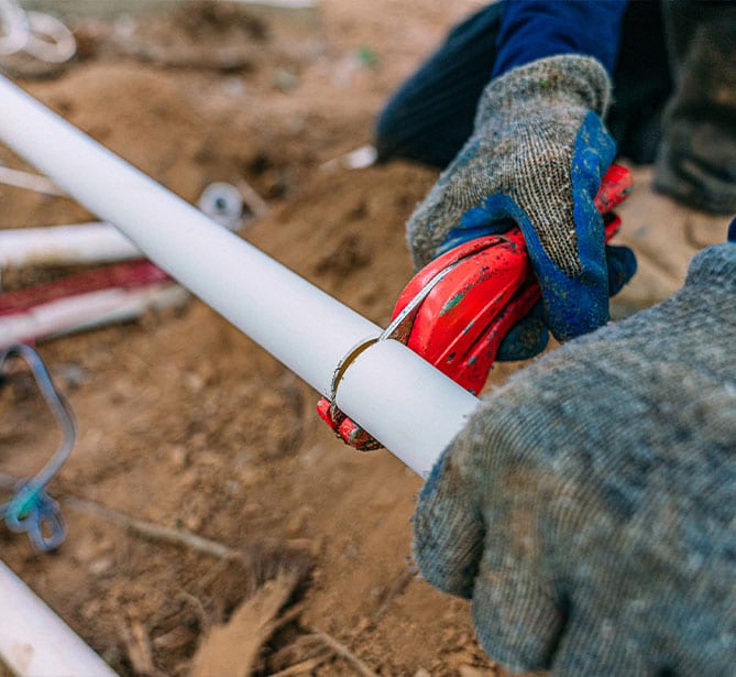 Gloved hands cutting a white PVC pipe outdoors with red tool.