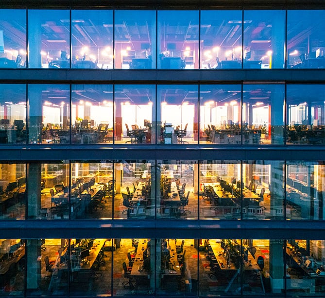 Glass-walled office building with multiple floors visible, illuminated workspaces inside.