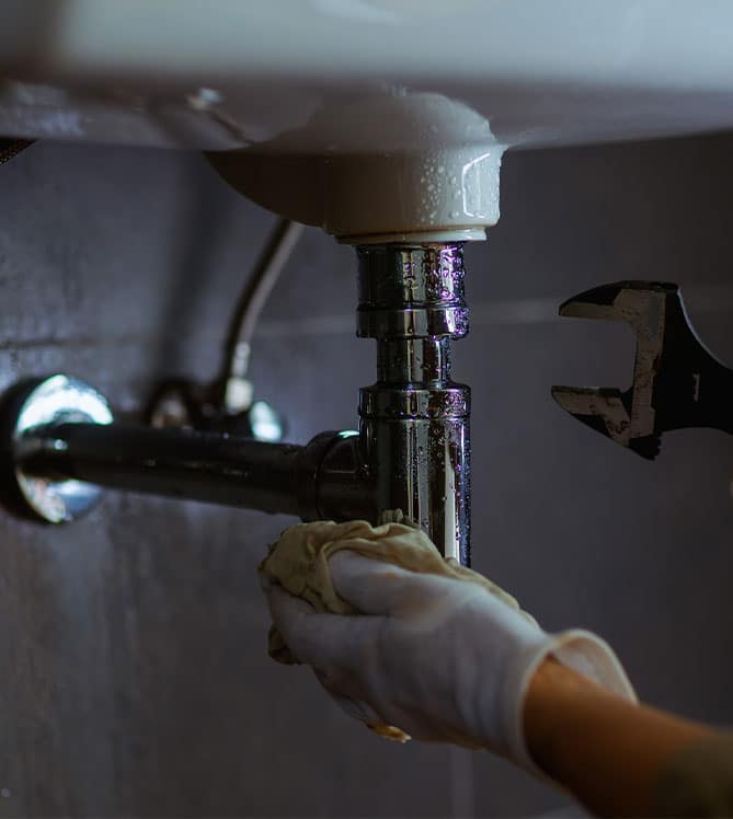 Plumber fixing a pipe under a sink with a wrench and gloves.