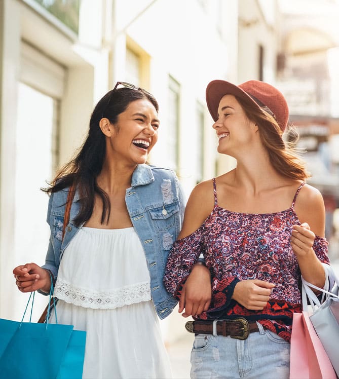 Two women laughing and shopping with bags on a sunny street.