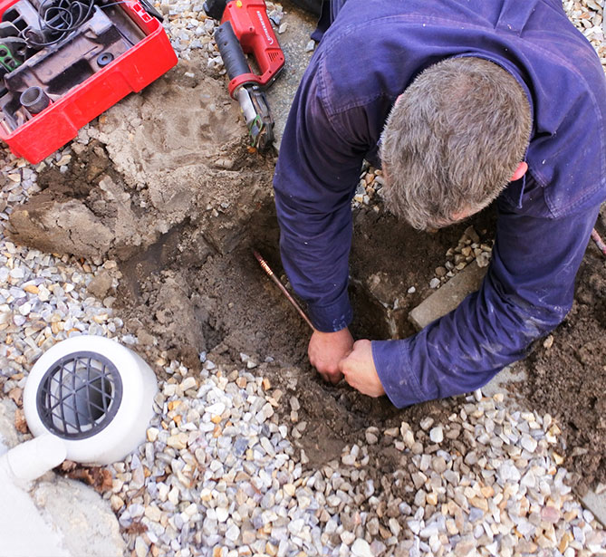 A person fixing pipes in the ground with tools on a gravel surface.